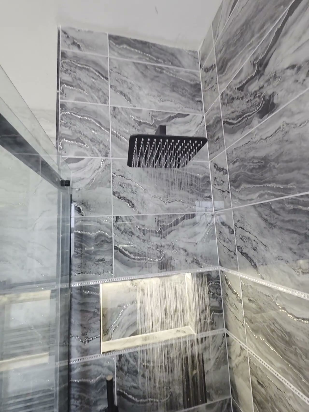 Walk-in shower with grey marble tiles, matte black rainfall shower and lit niche